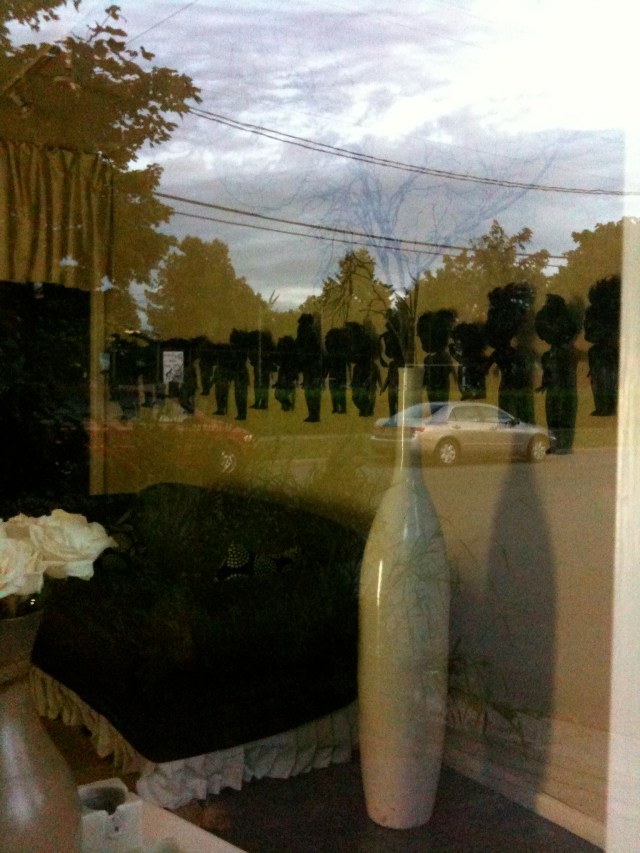 A photograph of a roomful of strangely assembled objects, seen through a window with reflections of the afternoon sky.